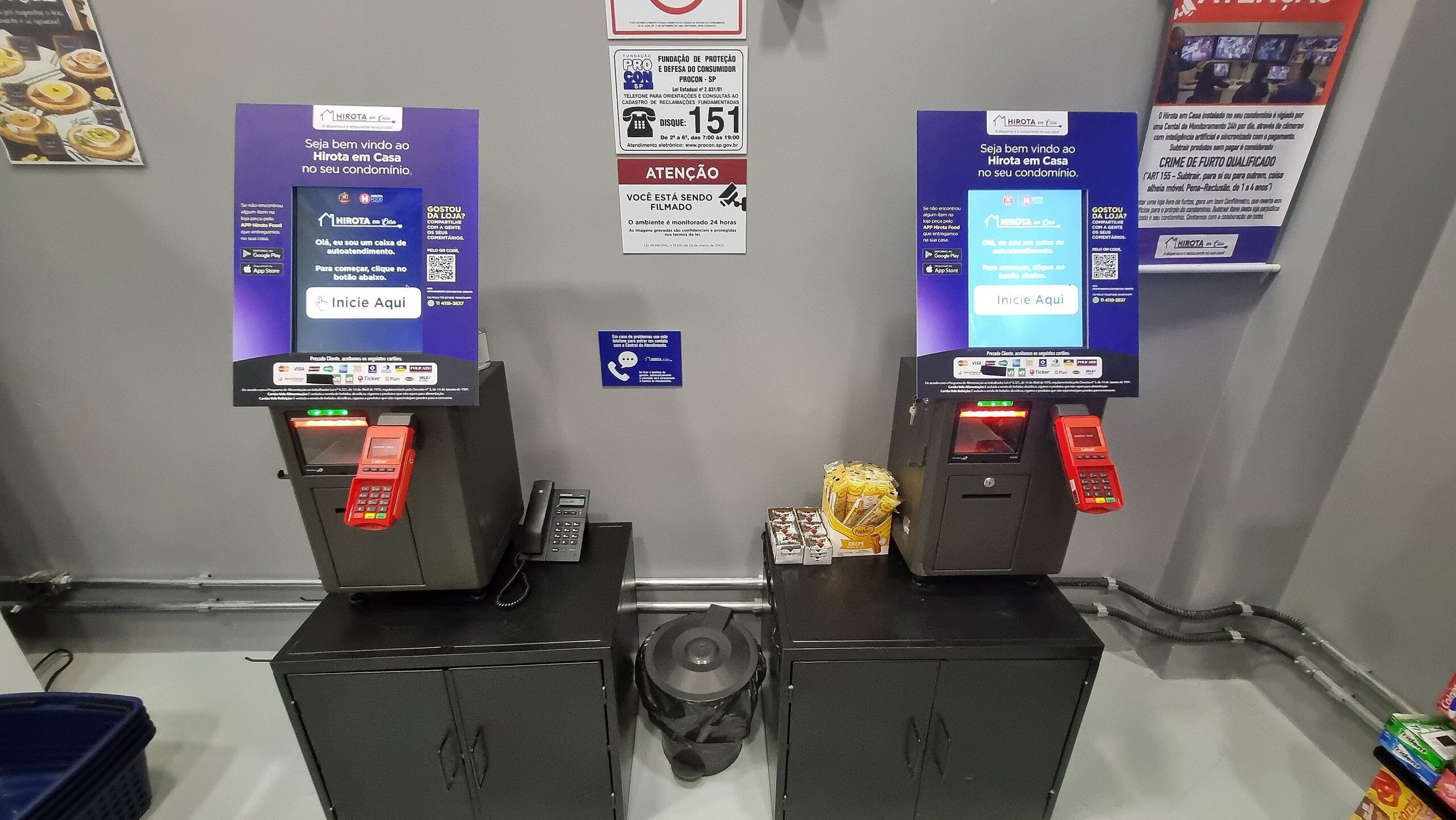 Dos quioscos de autoservicio en el vestíbulo de un hotel, con un cartel sobre cada uno que dice "HIROTA em Casa" y un código QR para iniciar una transacción.