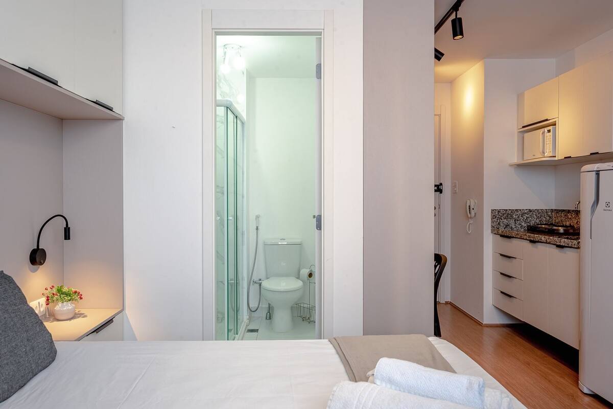 Moderni hotellihuone, jossa on sänky, yöpöytä ja pieni kylpyhuone, joka näkyy avoimesta ovesta.