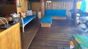 Una terraza de madera con un banco de madera y una mesa, un pequeño espacio de cocina y una zona de estar con cojines azules.