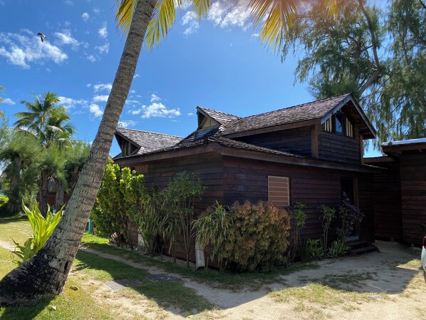 Ein Holzhaus mit geneigtem Dach, umgeben von Palmen und verschiedenen Sträuchern.
