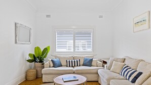 Ein Wohnzimmer mit einem beigen Sofa, einem runden Couchtisch, einer großen Topfpflanze und einem Fenster mit Jalousien.