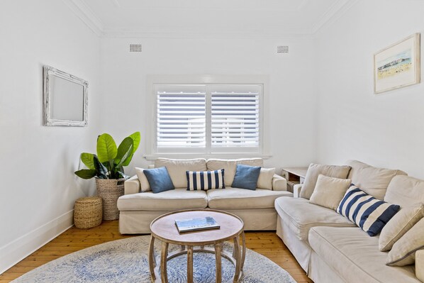A living room with a beige sofa, a round coffee table, a large potted plant, and a window with blinds.