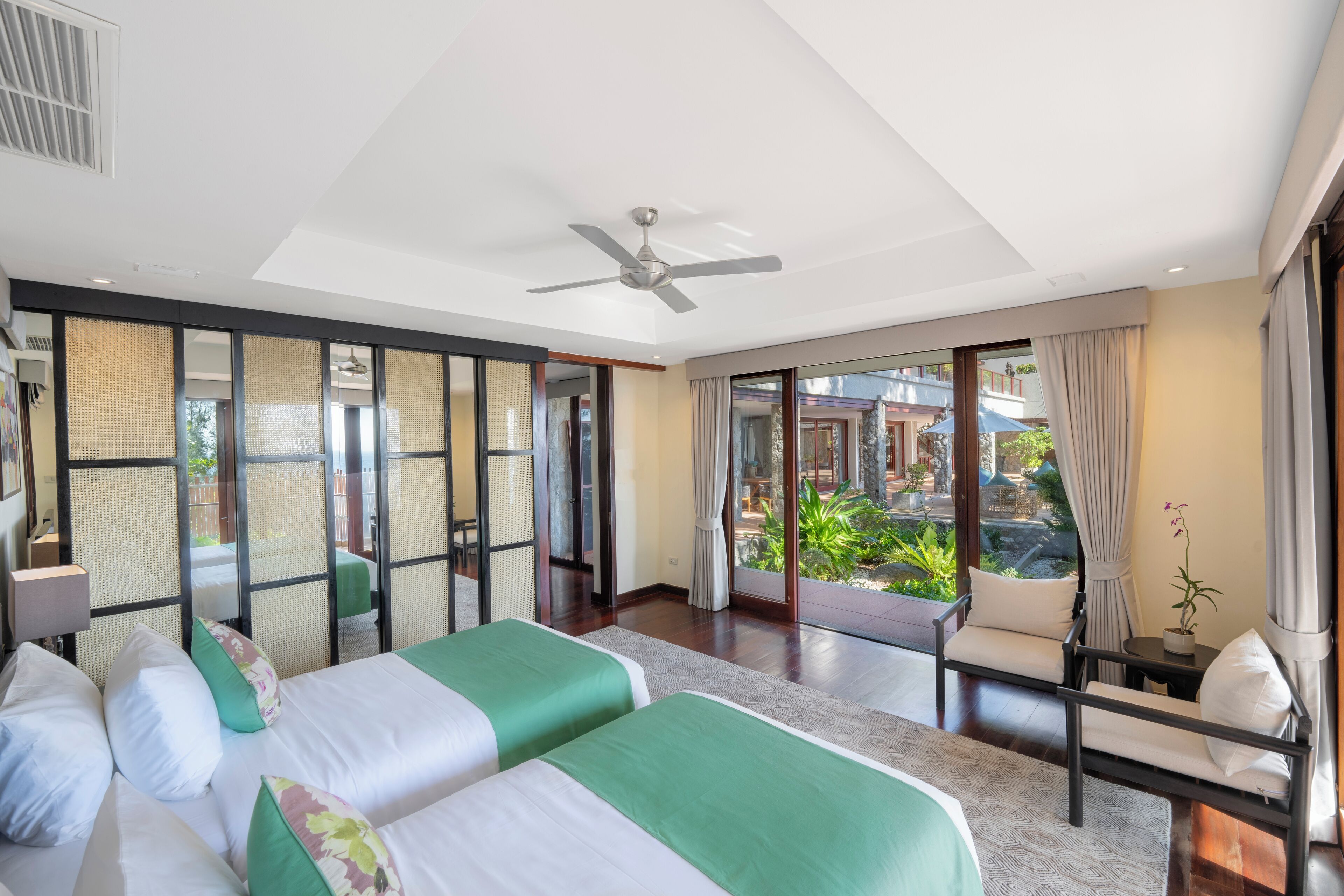A hotel room with two beds, a ceiling fan, a view of a garden, and a balcony.
