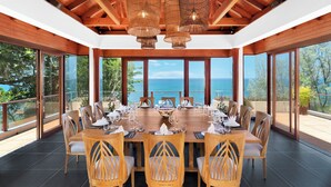 A dining area with a large table set for a meal, large windows with a view of the sea, and wooden ceiling beams. - Villa Horizon (Kamala)