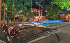 A poolside area with a lounge chair and a rose, a pool, and a building with outdoor lighting.