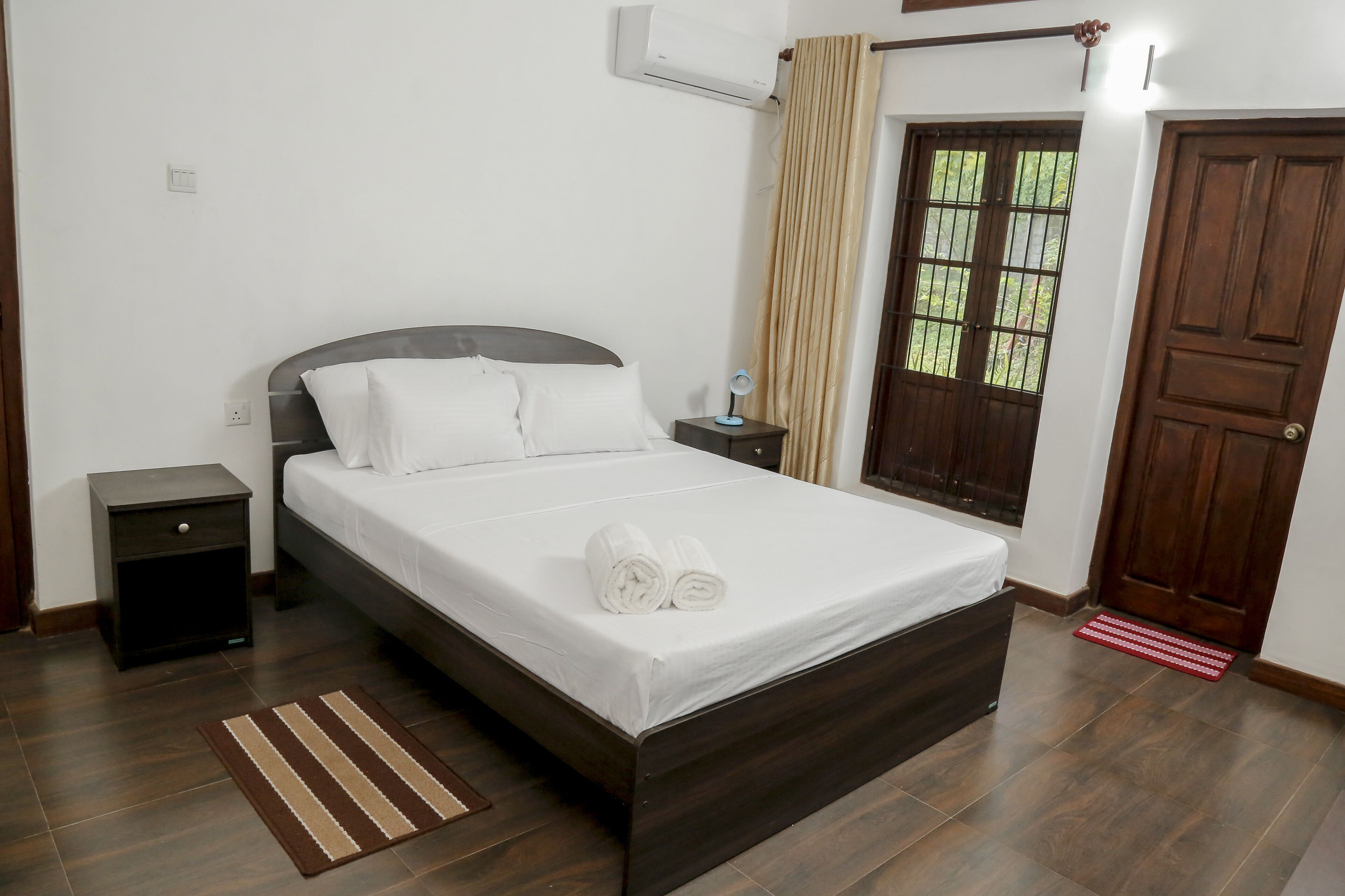 A neatly made bed with white linens and rolled towels, a bedside table with a lamp, a dark wooden nightstand, a red and white striped rug, a window with bars, and a wooden door.