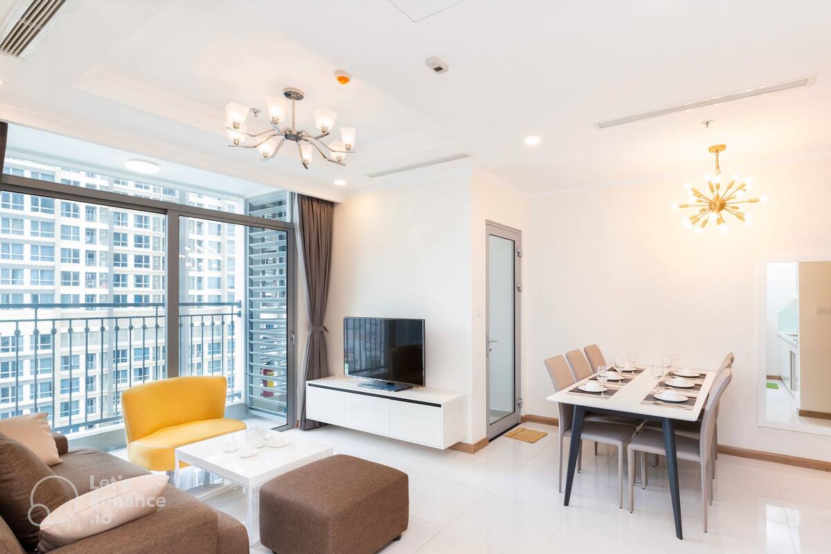 A modern living room with a sofa, a yellow chair, a dining table set for four, a TV on a white cabinet, and a large window with a city view.