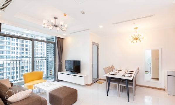 Uma sala de estar moderna com sofá, uma poltrona amarela, mesa de jantar para quatro pessoas, TV em um móvel branco e uma grande janela com vista para a cidade.