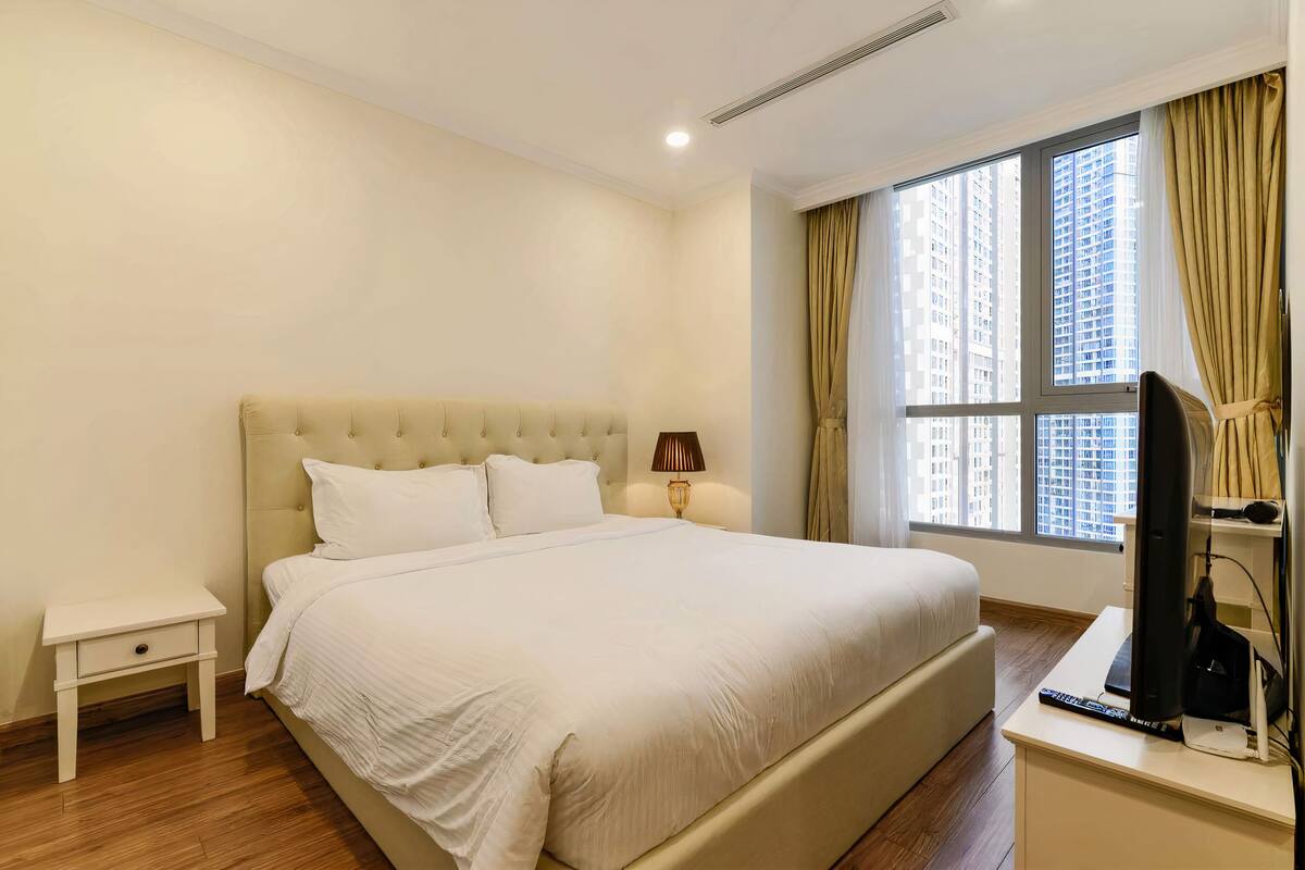 A bedroom with a large bed, a bedside table, a lamp, a TV, and a window with curtains.