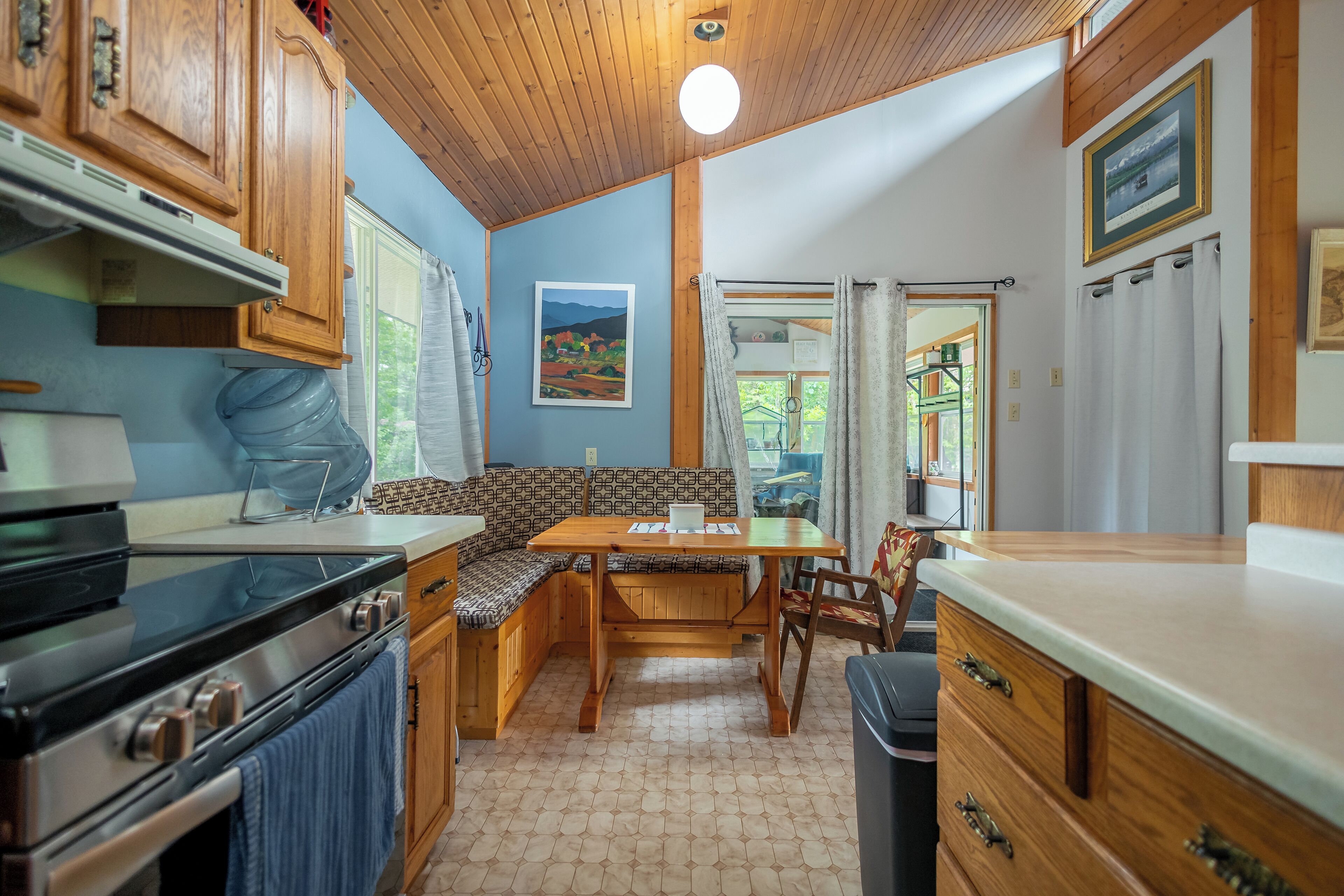A compact kitchen with wooden cabinets, a built-in stove, a dining table with benches, and a view of the outdoors through a window.