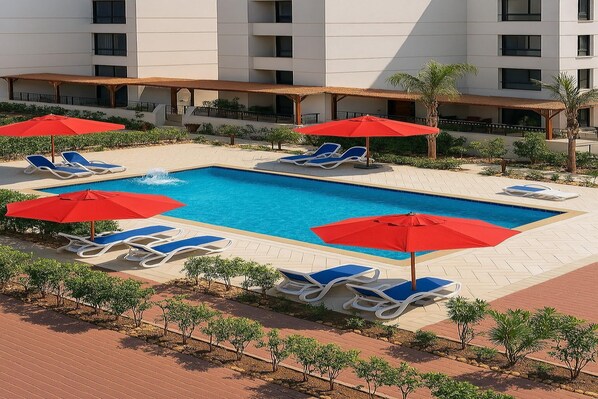 Zona de piscina hotelera con múltiples tumbonas y sombrillas rojas, rodeada de pequeños árboles y un edificio al fondo.