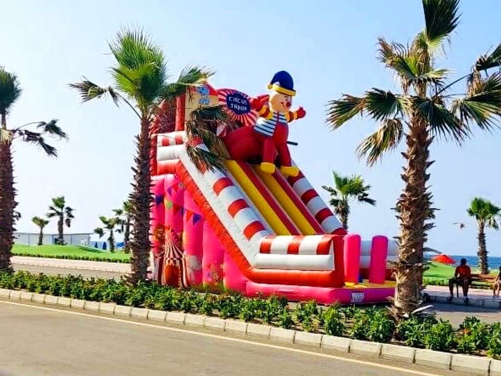 An inflatable slide structure with a circus theme, surrounded by palm trees and a clear sky.