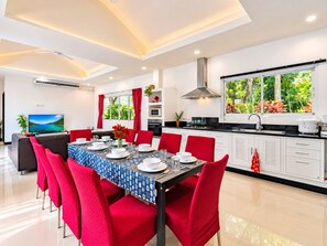 A modern dining area with a table set for six, red chairs, and a kitchen with white cabinets and a large window. - Phikun Private Pool Villa (Chalong)