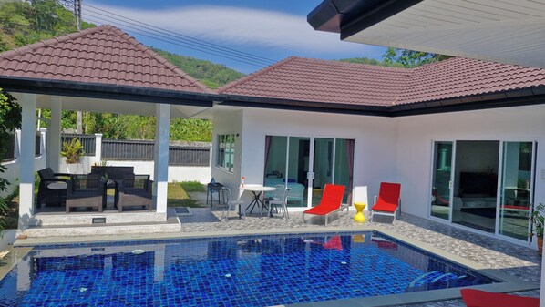 Zona de piscina con piscina azulejada, mobiliario exterior y un porche cubierto.