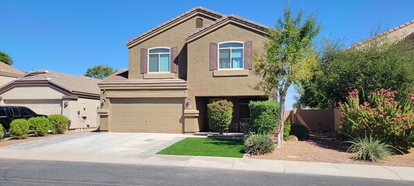 A two-story house with a garage, a well-maintained lawn, and various shrubs and flowers. - Charming 4 bed / 3 bath house, full amenities! (Sun City)