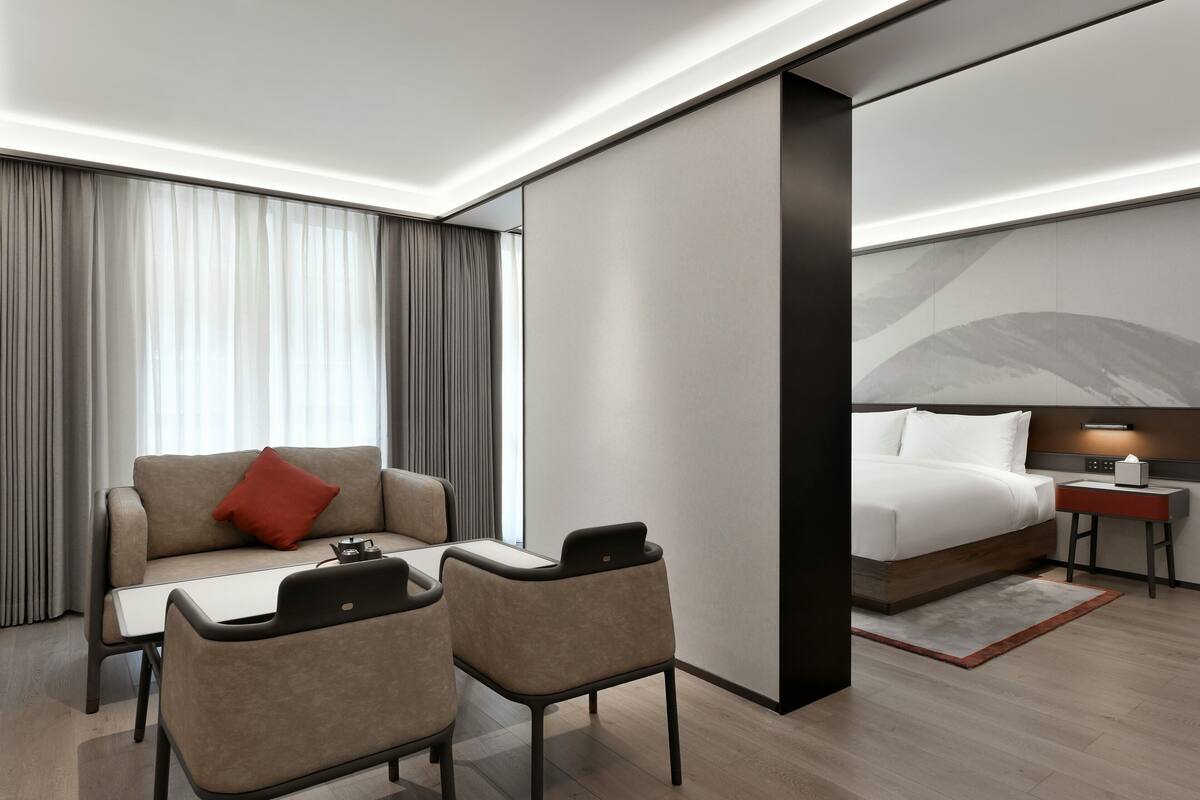 A modern hotel room with a sofa, two chairs, a small table, a bed, a nightstand, and a large window with curtains.