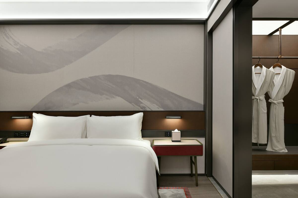 A modern hotel room with a large bed, bedside tables, a robe rack, and a wall-mounted artwork.