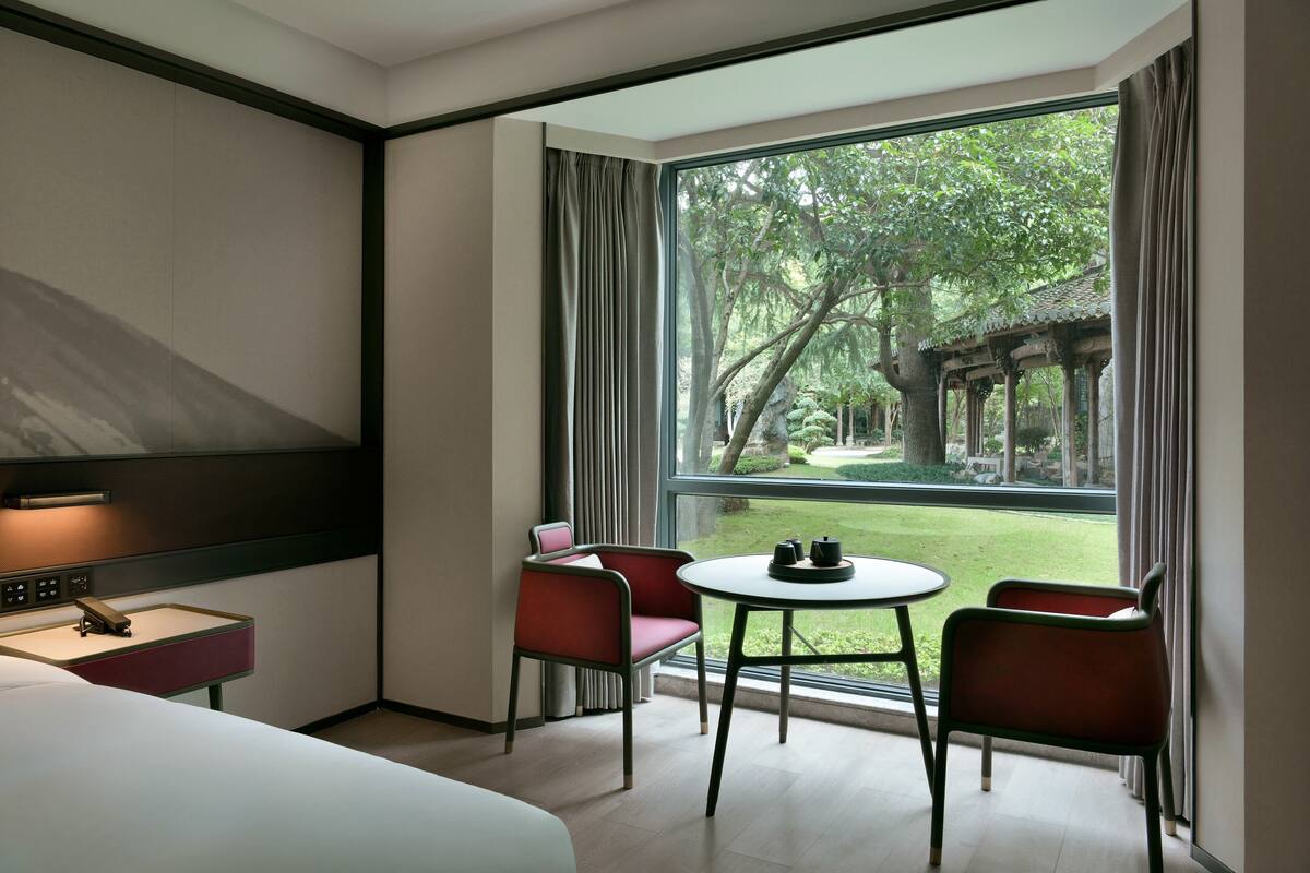 A hotel room with a bed, a desk with a lamp, a small table with chairs, and a large window offering a view of a garden and trees.