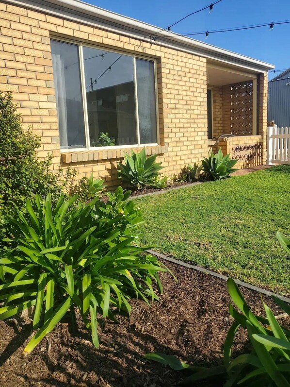 A residential house with a brick facade, large windows, and a well-maintained lawn with various plants.
