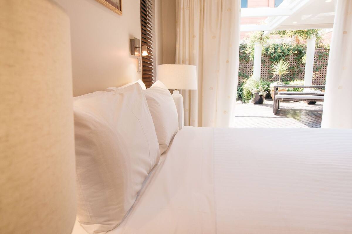 A hotel room with a large bed, a nightstand with a lamp, and a view of an outdoor patio with a bench and plants.