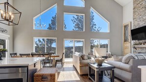 A spacious living area with a large dining table, a sofa, and a fireplace. There are multiple windows providing a view of the outdoors. - Mountain Dream, New Modern Home with Hot Tub and Spectacular Views (Divide)