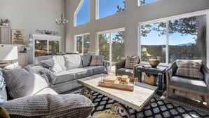 A spacious living room with a grey sofa set, a coffee table, and a view of the outdoors through large windows. - Mountain Dream, New Modern Home with Hot Tub and Spectacular Views (Divide)