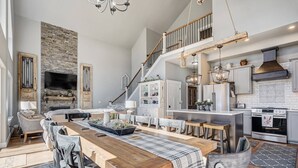 A spacious living area with a wooden dining table, comfortable seating, a stone accent wall, a TV, and a kitchen with modern appliances. - Mountain Dream, New Modern Home with Hot Tub and Spectacular Views (Divide)