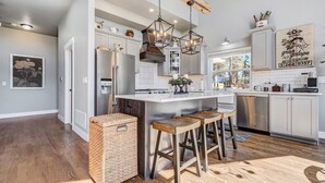 A modern kitchen with a central island, bar stools, a hanging light fixture, and a rustic ceiling beam. - Mountain Dream, New Modern Home with Hot Tub and Spectacular Views (Divide)
