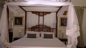Una cama con dosel y cuatro columnas, un cabecero de madera, dos mesitas de noche con lámparas y cuadros enmarcados en las paredes.