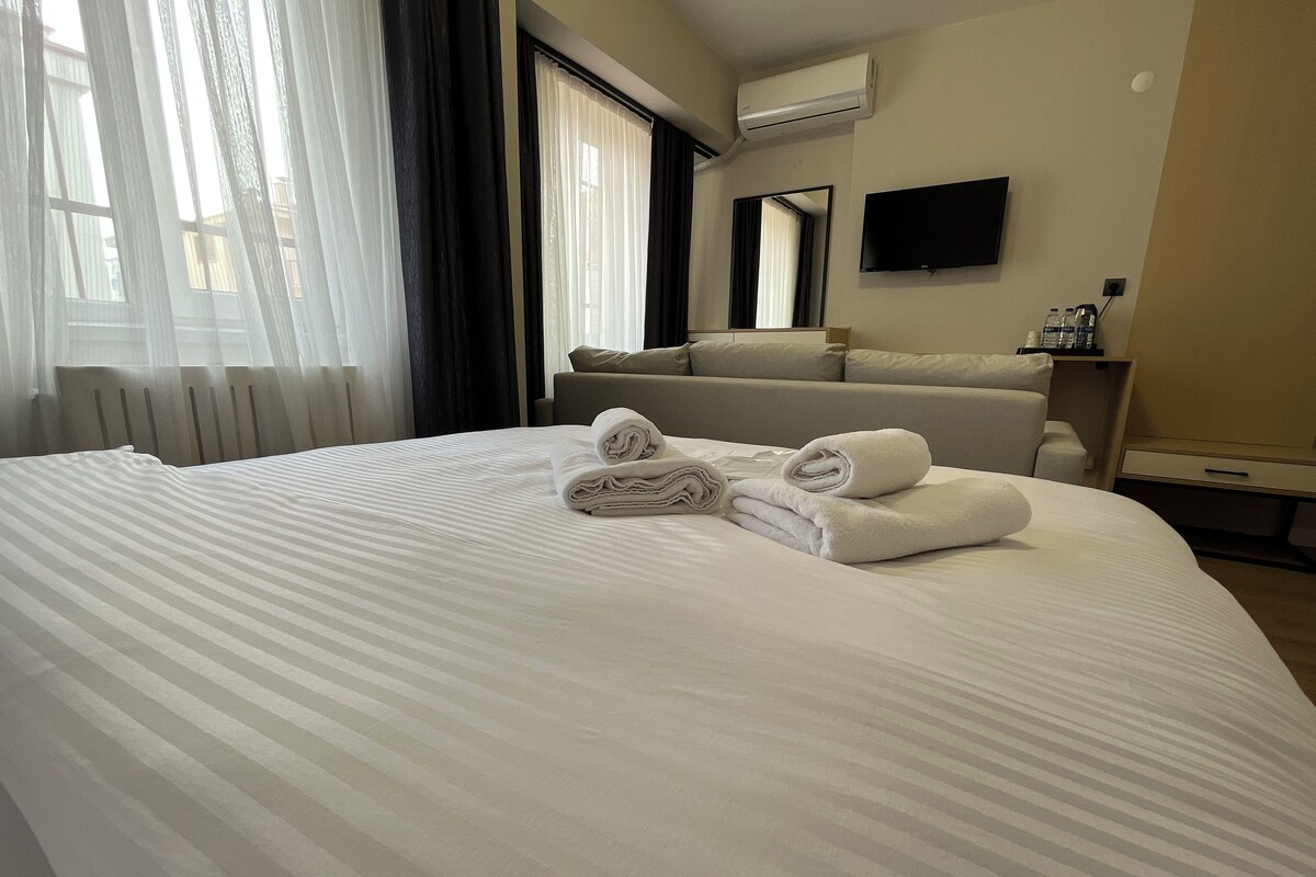 A neatly made bed with rolled towels, a television mounted on the wall, and a window with curtains.