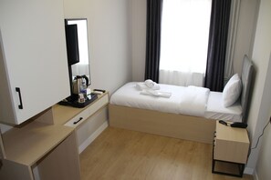Habitación de hotel pequeña con una cama individual, una mesita de noche con lámpara, un escritorio con silla, un espejo y una ventana con cortinas.