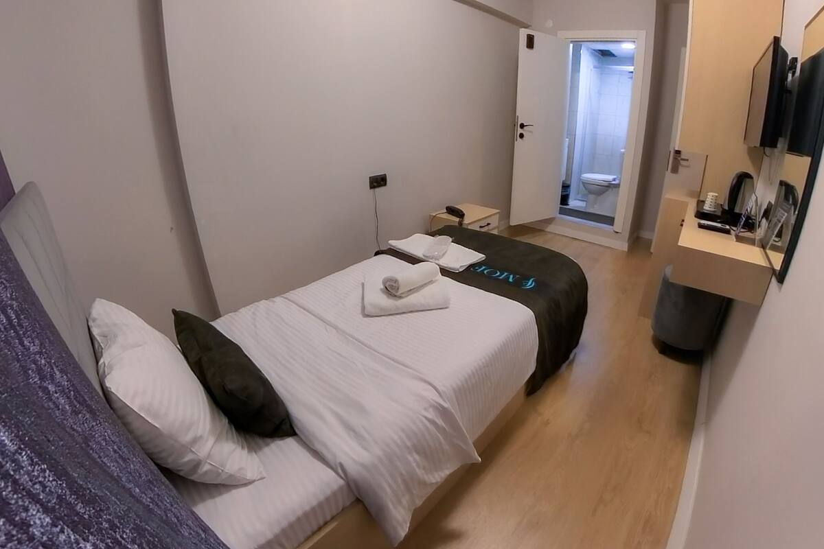 Ein kleines, modernes Hotelzimmer mit einem Einzelbett, einem Nachttisch, einem Spiegel und einem durchgegangenen Türrahmen einsehbaren Badezimmer.