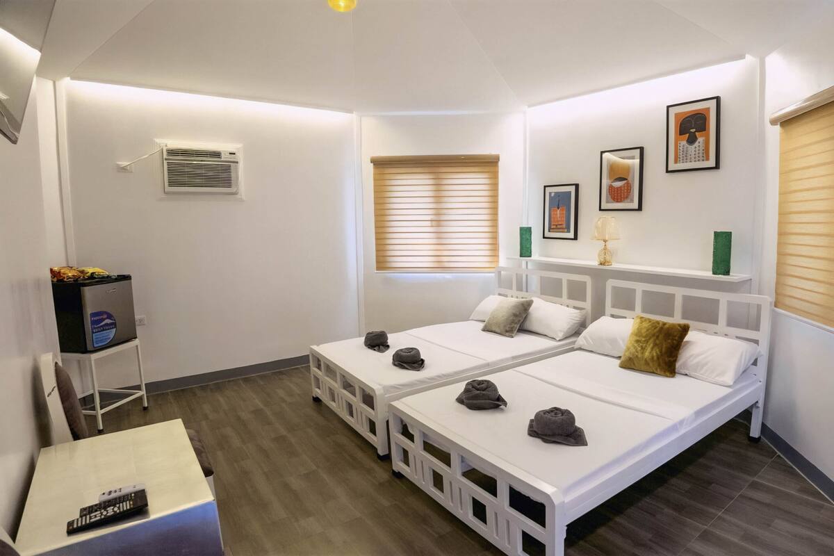 Ein Hotelzimmer mit zwei Einzelbetten, einem kleinen Tisch mit Fernbedienung, einer Minikühlschrank und einem Fenster mit Jalousien.