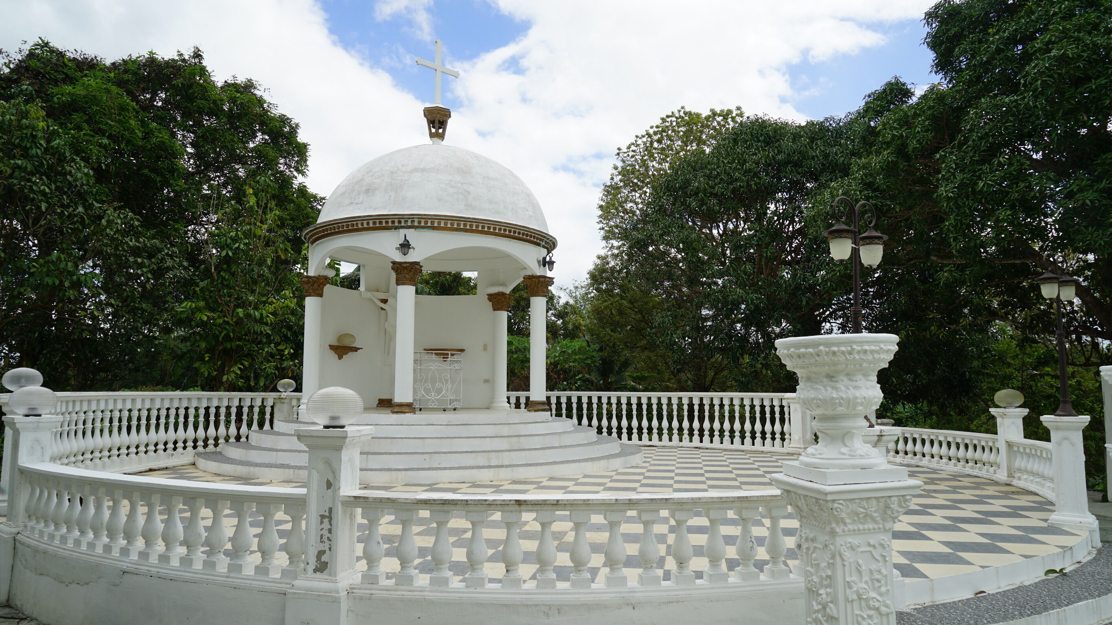 A white gazebo with a dome roof and a cross on top, surrounded by a checkered floor and white balustrades.