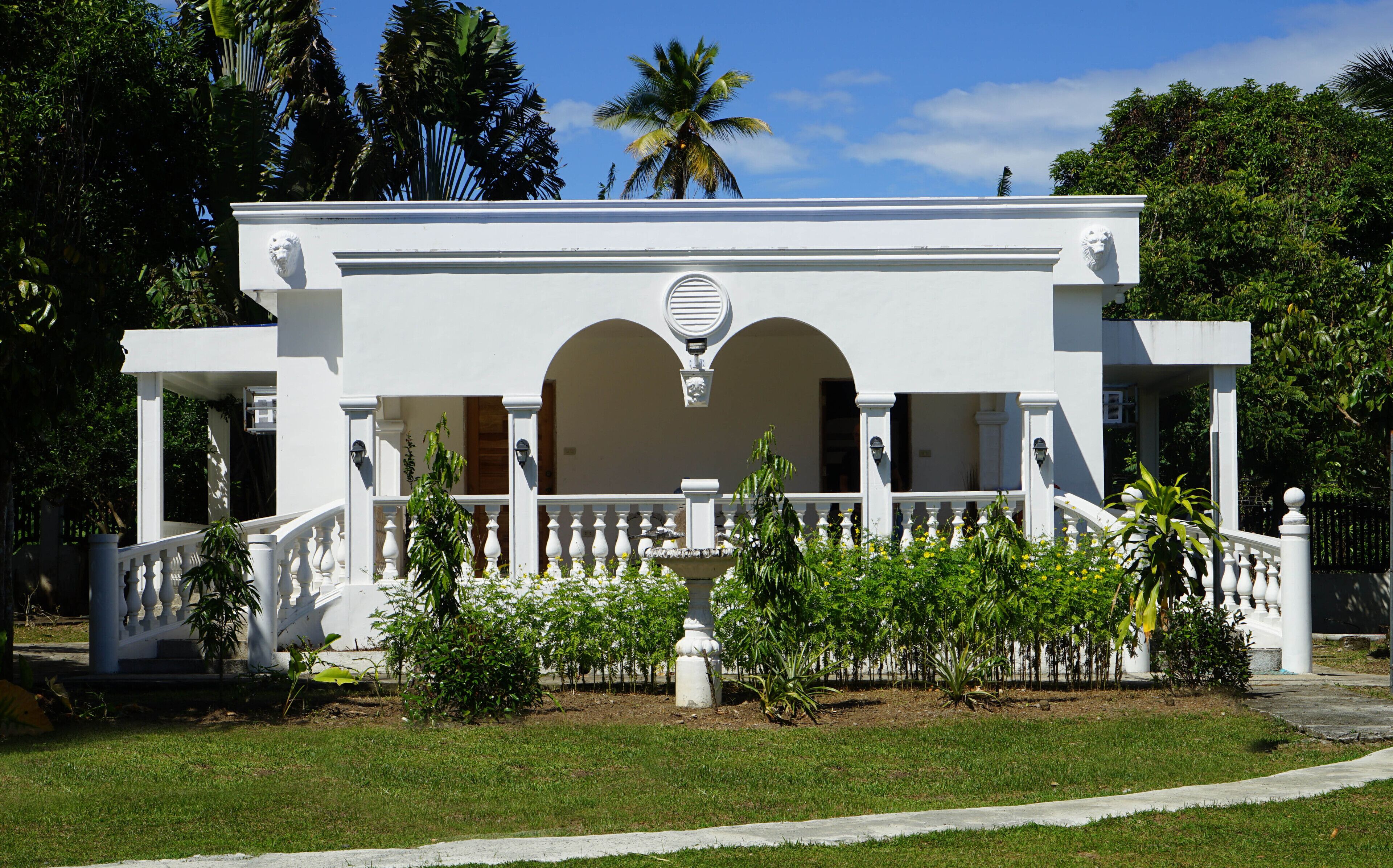 Un bâtiment blanc doté d’une arche centrale, d’un balcon blanc orné de balustrades et d’un jardin à l’avant.