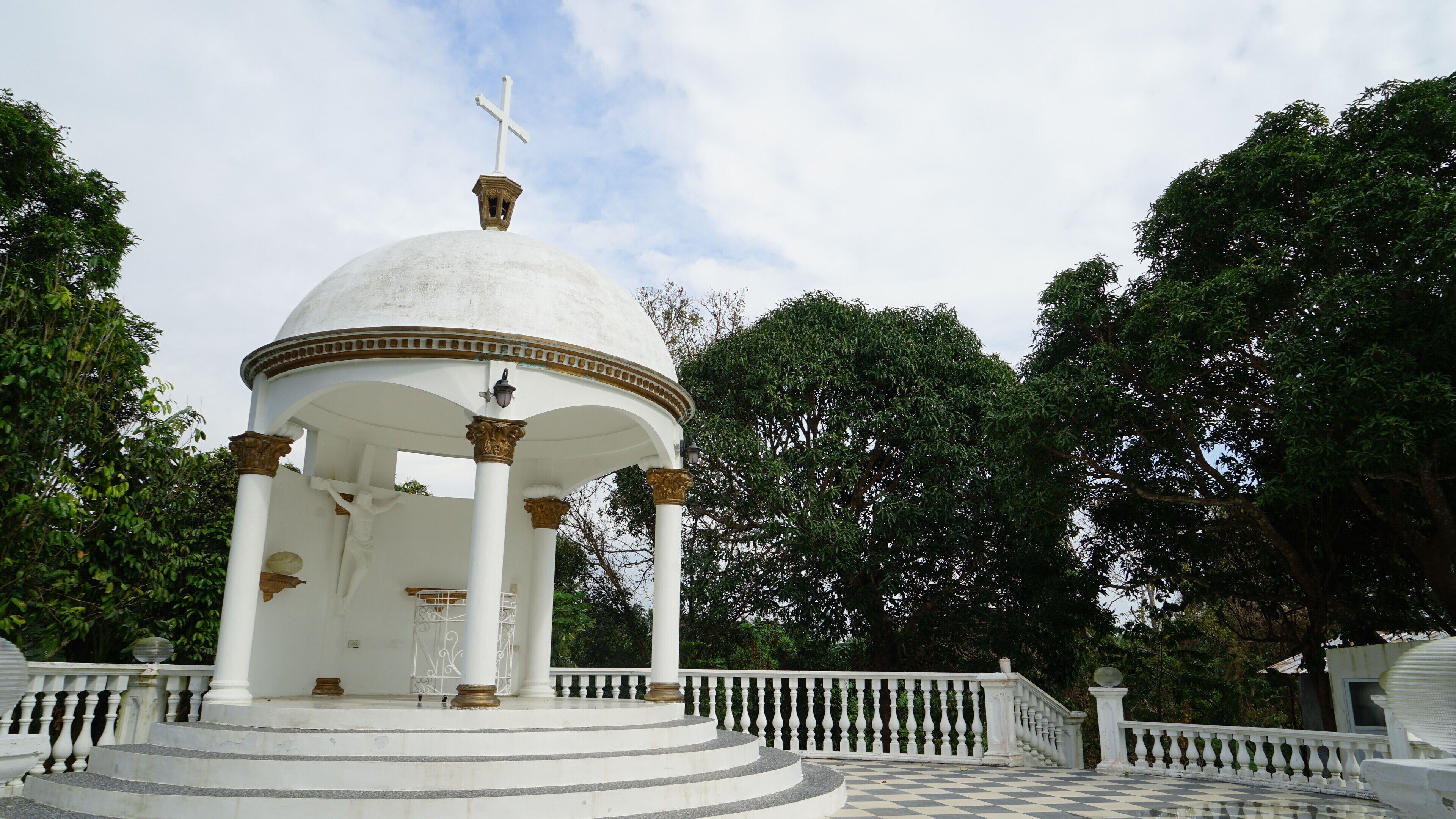 A white gazebo with a dome and a cross on top, surrounded by a checkered floor and white balustrade.