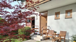 A modern outdoor seating area with a wooden table and chairs, a small garden with a pathway, and a building entrance with a wooden door. - Ferienwohnung 40m² mit Terrasse, bis 3 Personen, Separates Schlafzimmer (Grassau)