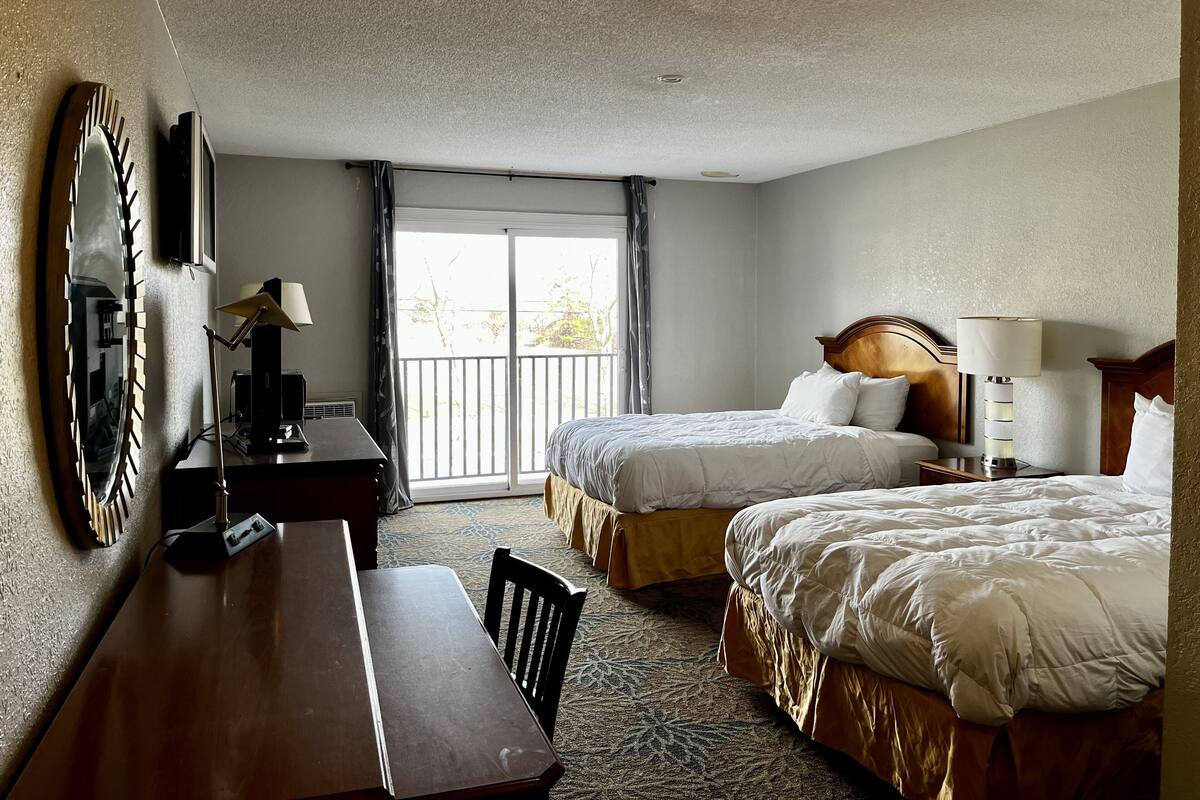 Habitación de hotel con dos camas, un escritorio, una silla, una lámpara y vistas al exterior.