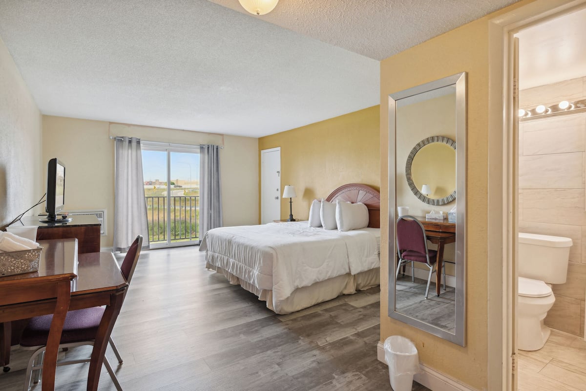 Habitación de hotel con cama, escritorio y silla, baño con inodoro y un espejo grande.