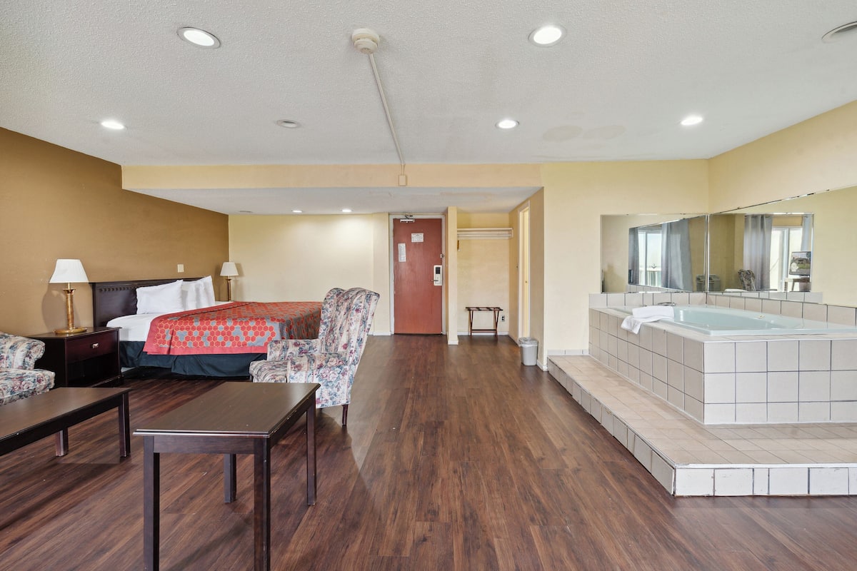 Habitación de hotel con cama, dos sillas, una mesa, escritorio, espejo, bañera y puerta que da a otra habitación.