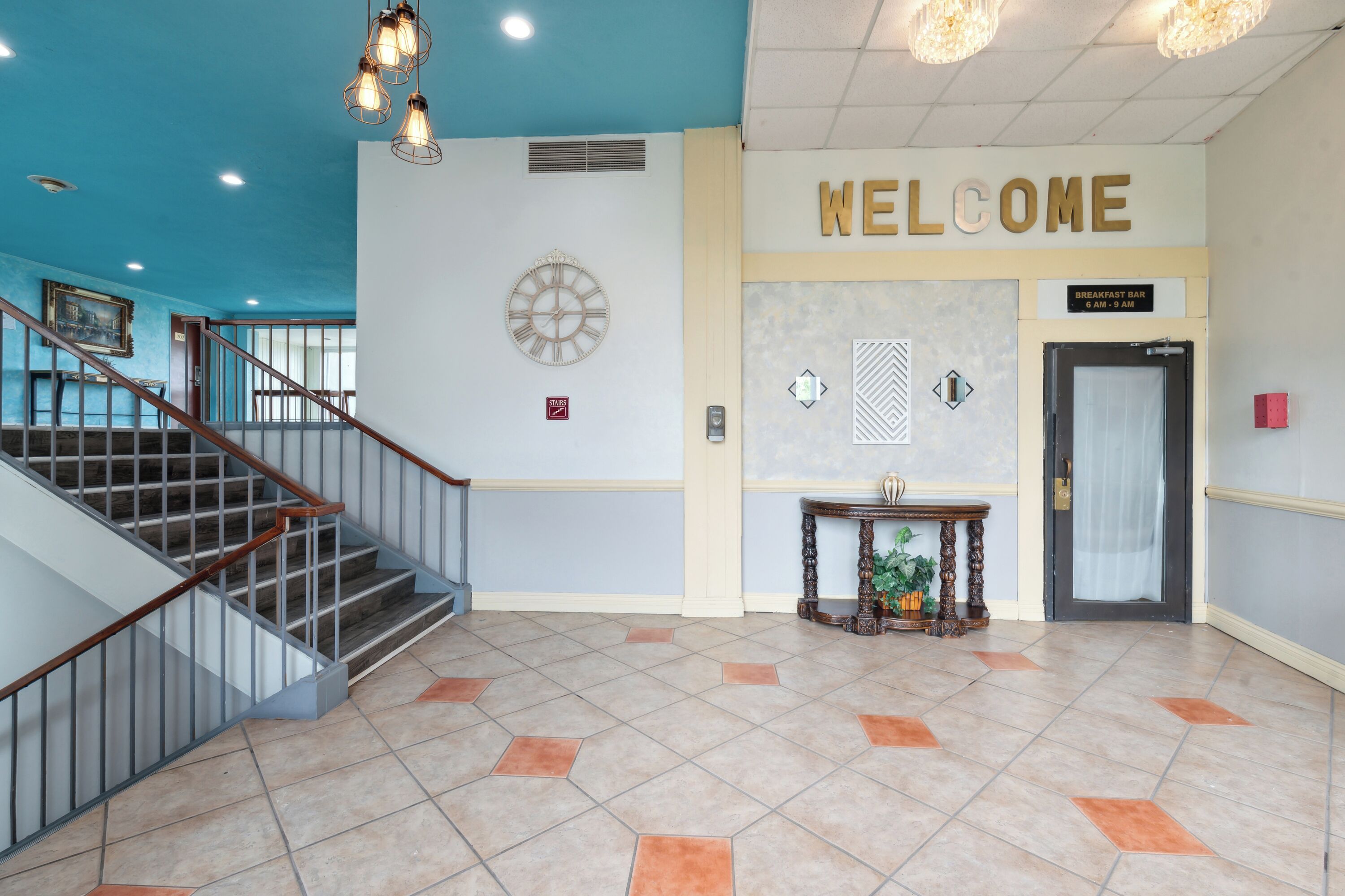 A hotel lobby with a staircase, a welcome sign, a clock, and a breakfast bar.