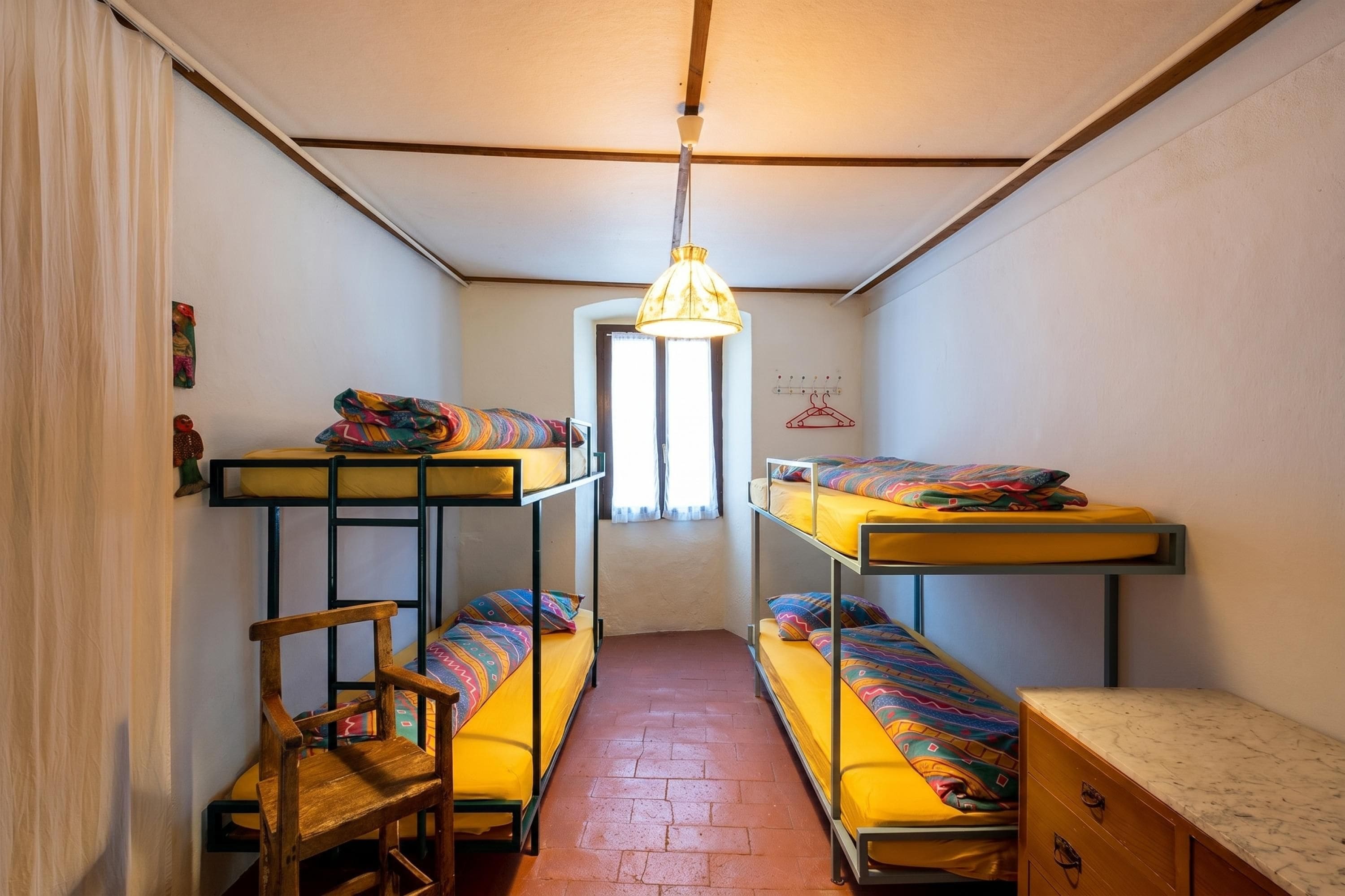 A narrow dormitory room with bunk beds, a wooden chair, a desk, and a hanging light fixture.