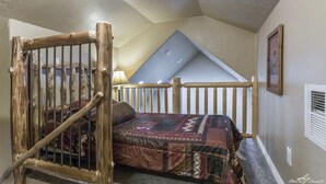 A loft bedroom with a sloped ceiling, a wooden ladder, a bed with a patterned bedspread, and a framed picture on the wall.
