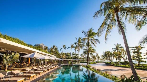 A poolside area with lounge chairs and umbrellas, surrounded by palm trees and a clear sky.