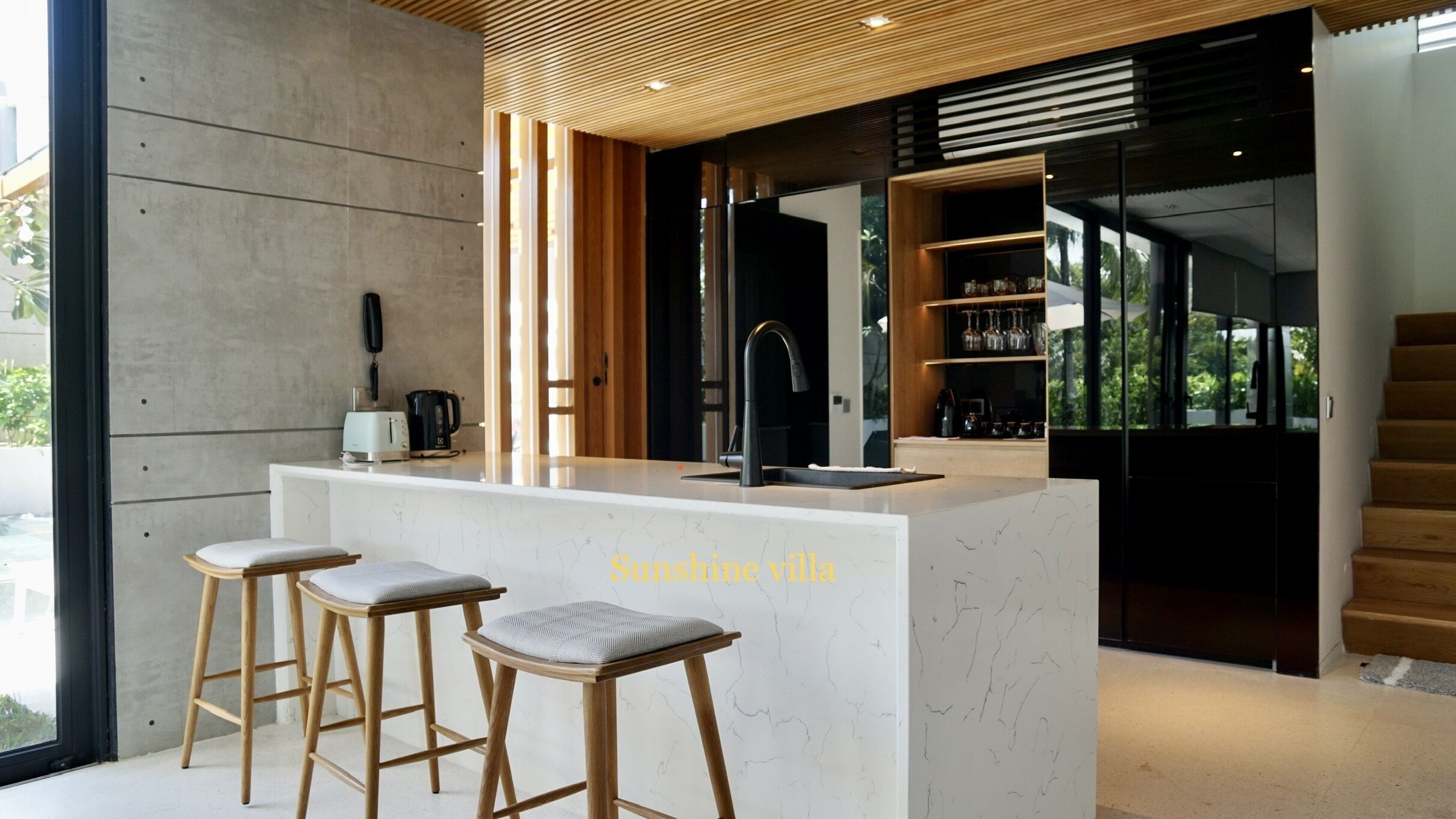 A modern kitchen with a marble countertop, wooden stools, and a built-in coffee machine.