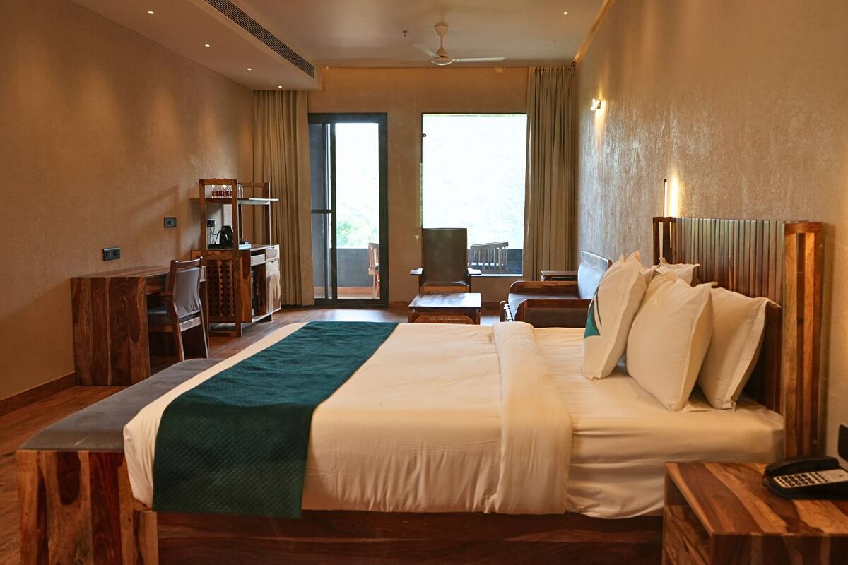 A hotel room with a large bed, a desk, a chair, and a view of the outdoors through the windows.