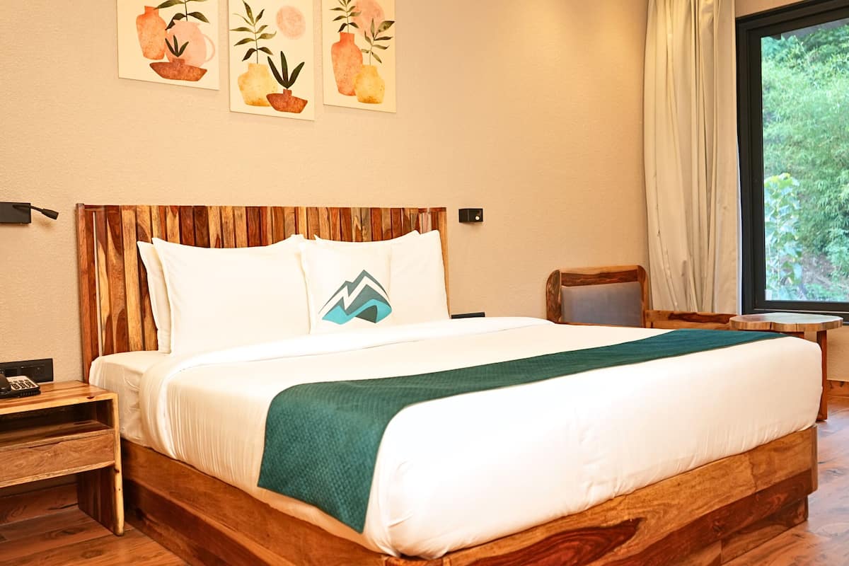A wooden bed with white bedding and a green throw, a wooden nightstand with a phone, and a window with a view of greenery.