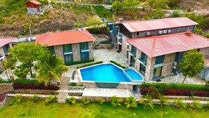 A modern resort with a central swimming pool, stone and glass architecture, and lush landscaping. - The Divine Hills Resort | Suites with Private Pool | Near IIM Udaipur (Udaipur)
