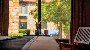 A hotel room with a bed, a wooden chair, a window with a view of a stone building and greenery. - The Divine Hills Resort | Suites with Private Pool | Near IIM Udaipur (Udaipur)