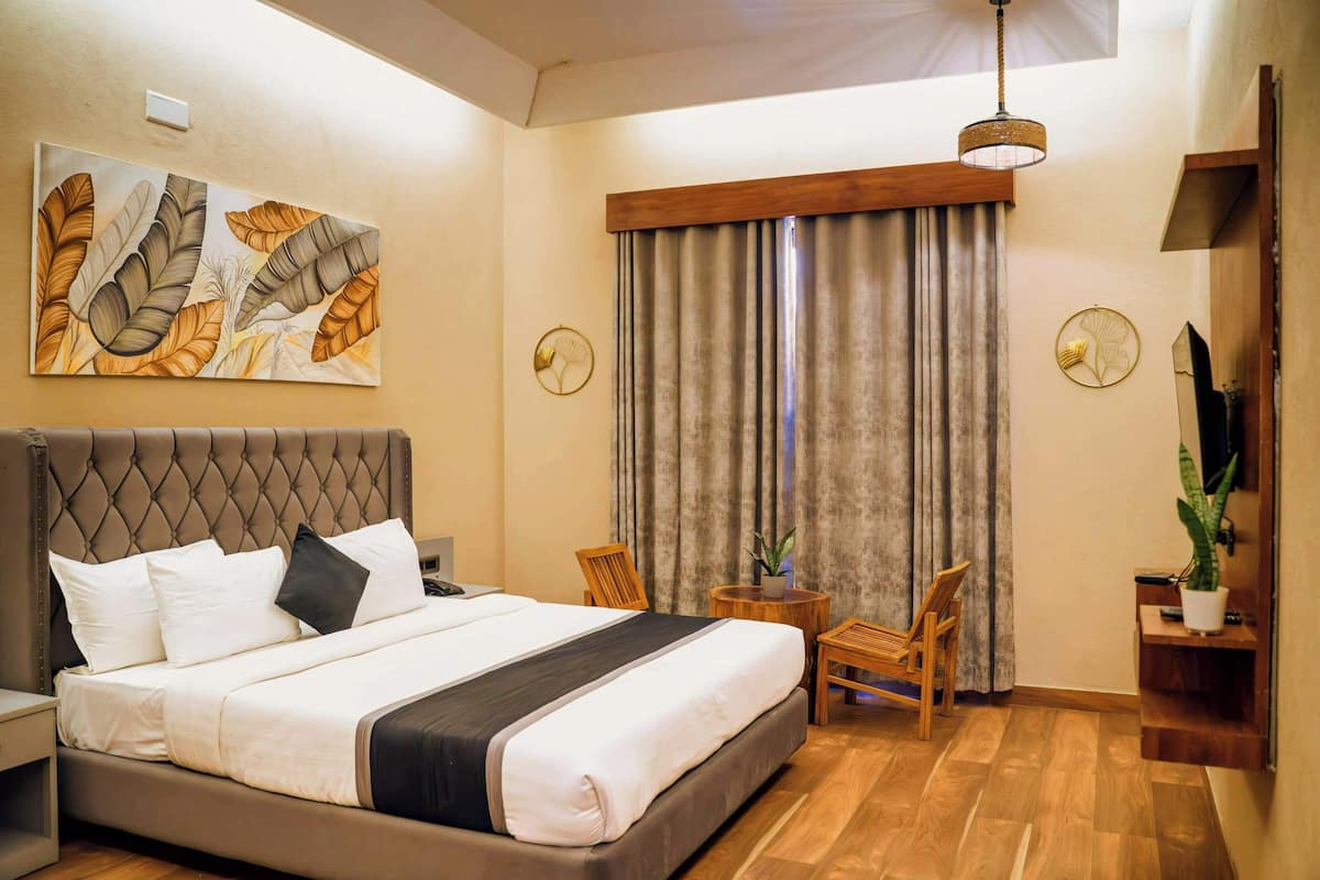 A hotel room with a large bed, a wooden headboard, a small table with a chair, a potted plant, and a large window with curtains.